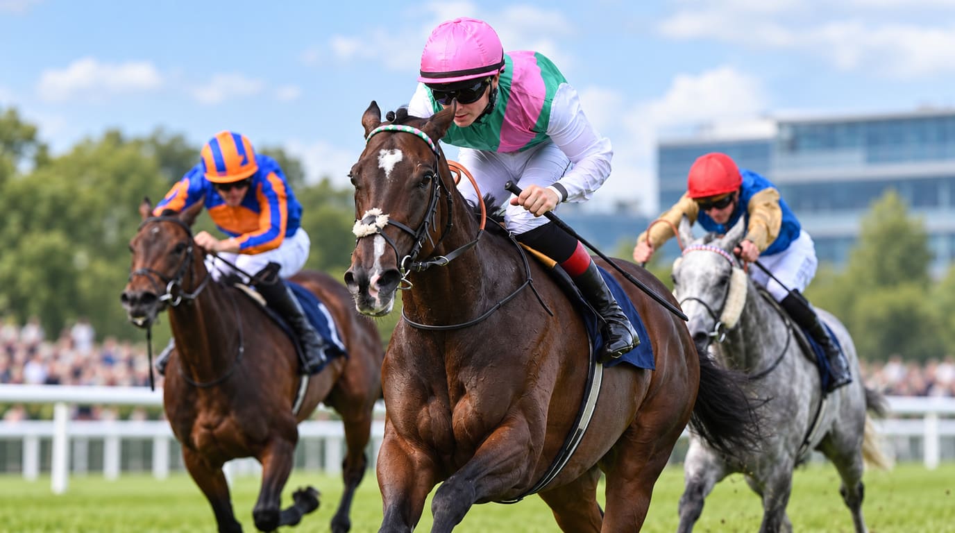 Bay filly with a white blaze and a jockey in pink and green silks leads a bay and a grey horse down a turf straight in front of a grandstand.