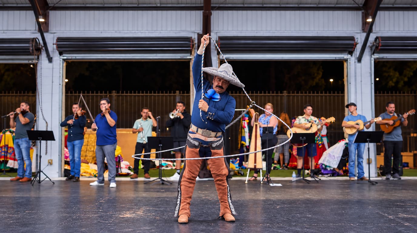 A charro in a navy shirt and suede chaps spins a large rope loop around himself on stage while a mariachi band plays behind him.