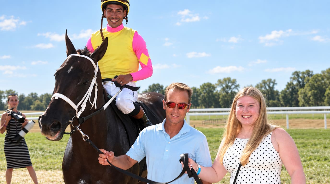 Un jinete sonriente con chaquetilla amarilla y rosa monta un caballo de carreras castaño oscuro, guiado por un hombre y una mujer en un día soleado.