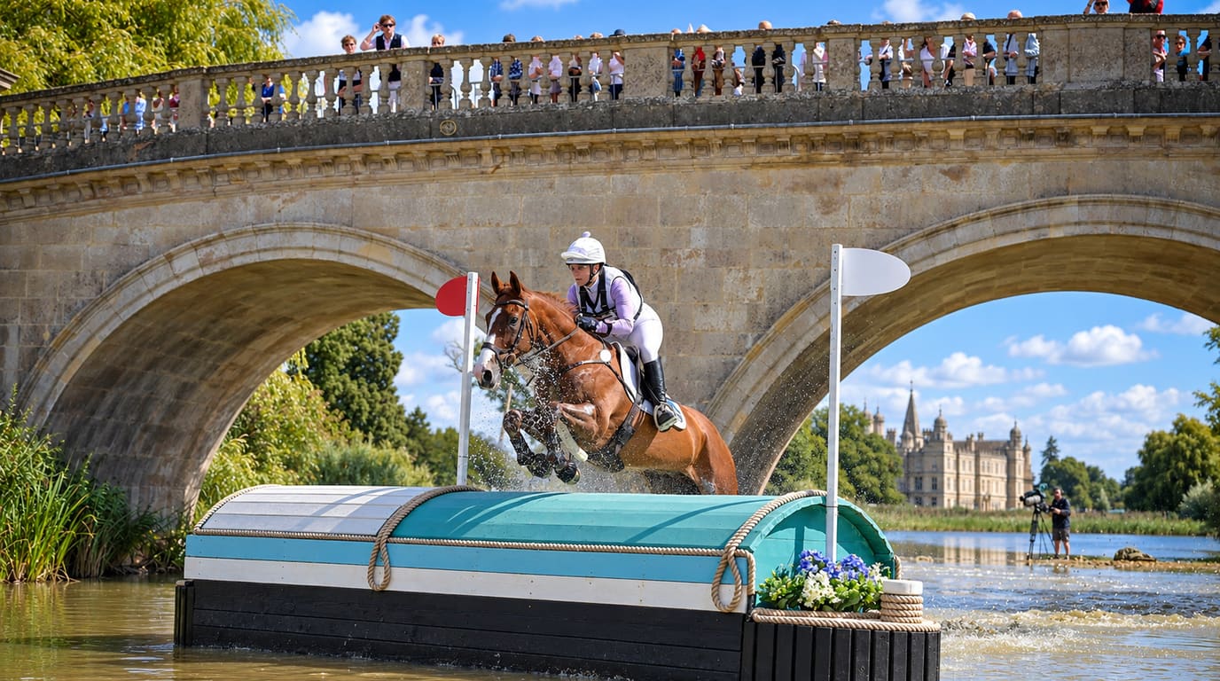 An eventing rider on a chestnut horse leaps a boat-shaped water fence beneath a stone bridge, with a grand country house in the distance.